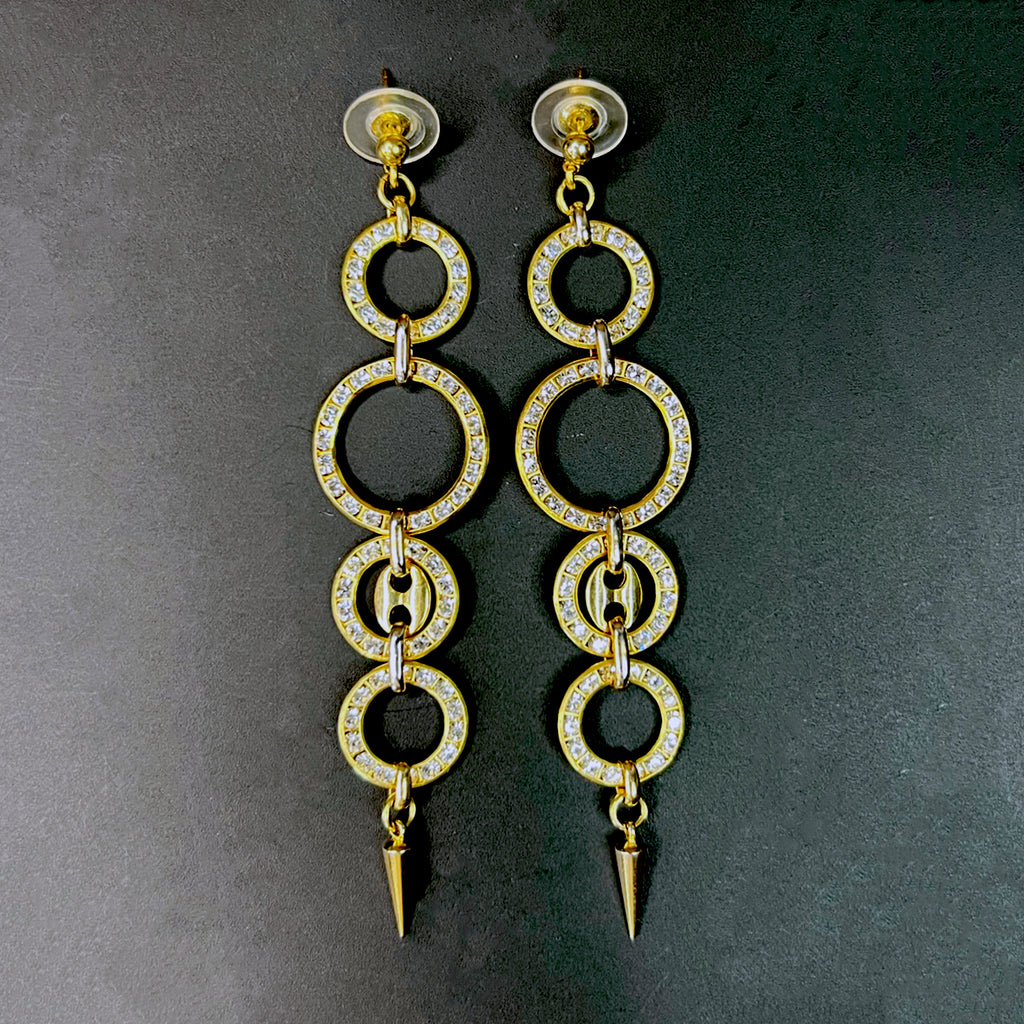 Flat lay of a matching pair of gold chandelier earrings with rhinestone-encrusted hoops and pointed spikes, arranged symmetrically on a dark surface.