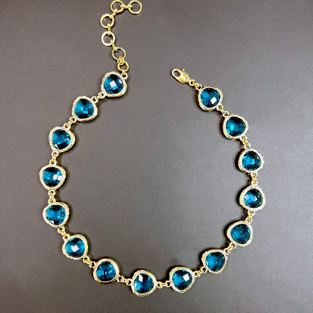 Flat lay of a gold-plated choker necklace featuring round aqua blue faceted crystals in textured bezel settings, arranged on a black background.