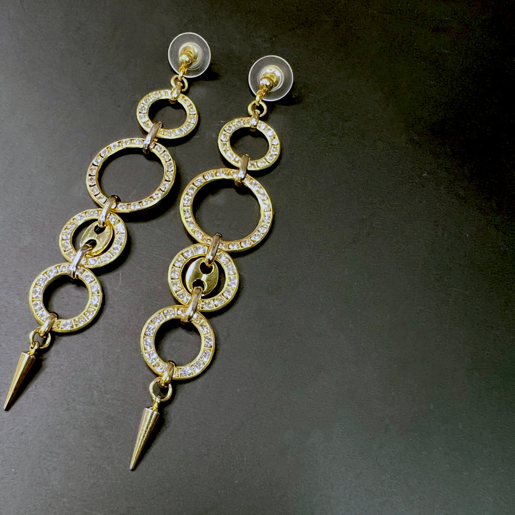 Close-up of long gold dangle earrings featuring five interlinked circular hoops encrusted with rhinestones, ending in a sleek pointed spike, displayed on a dark surface.