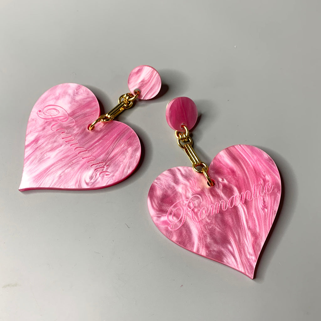 Oversized pink acrylic heart earrings with "Romantic" engraving and gold chain detailing on black ear display