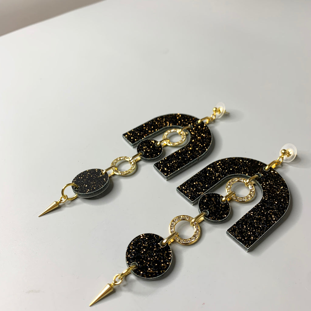 Angled image of the black and gold chandelier earrings with shimmering crystals and bold silhouette design on light gray surface