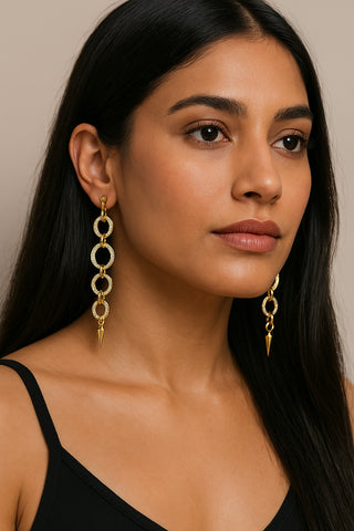 Woman wearing long gold chandelier earrings with rhinestone-studded interlocking circles and a pointed spike at the bottom, styled with a sleek black tank top.