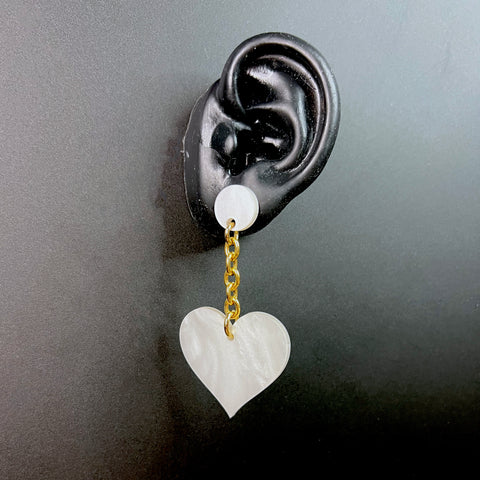 White heart-shaped acrylic drop earring hanging from a gold chain on a black ear display – statement jewelry.