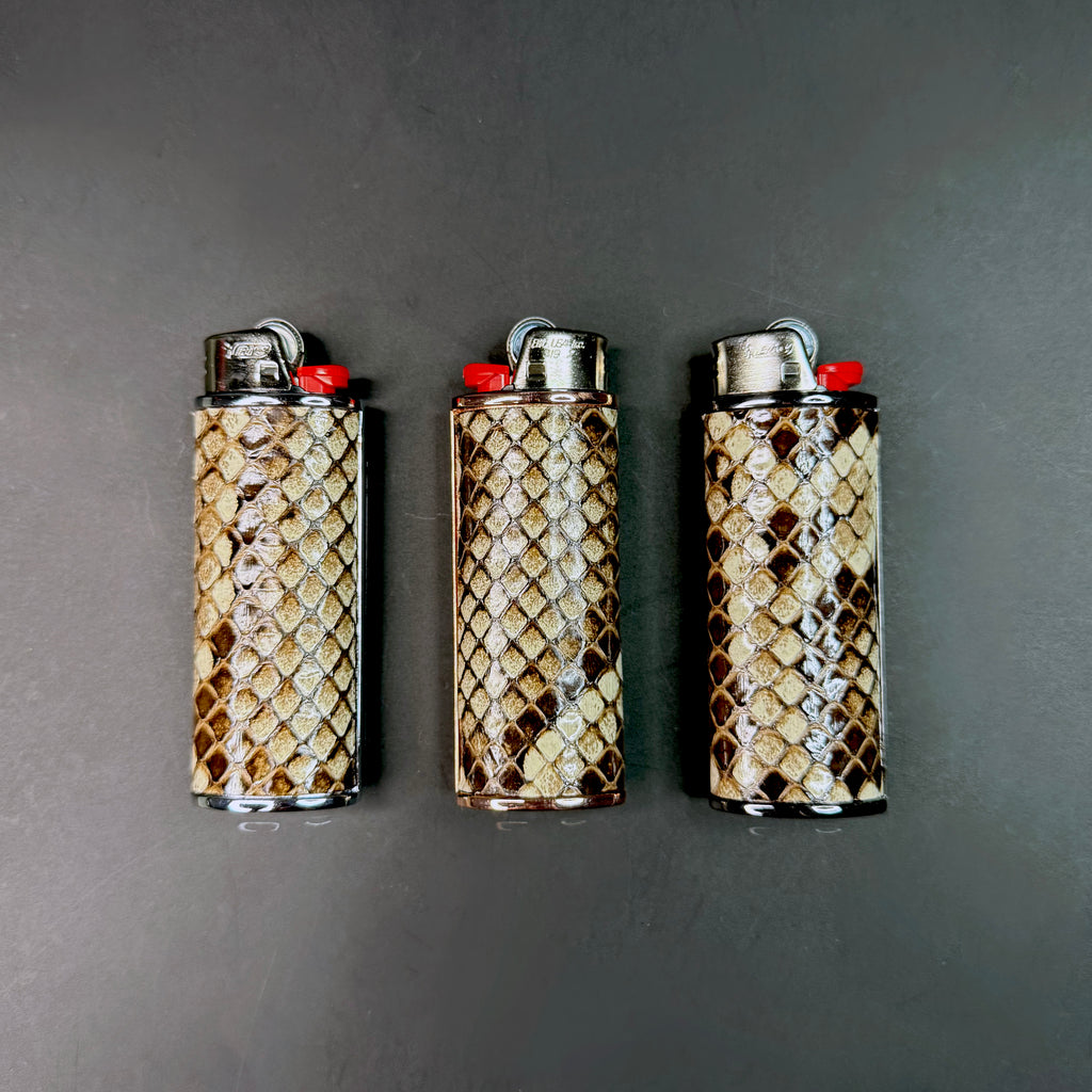 Three Burmese natural python-wrapped lighters displayed side by side on a dark background, each featuring unique scale patterns and metallic bases in rose gold, silver, and gunmetal.