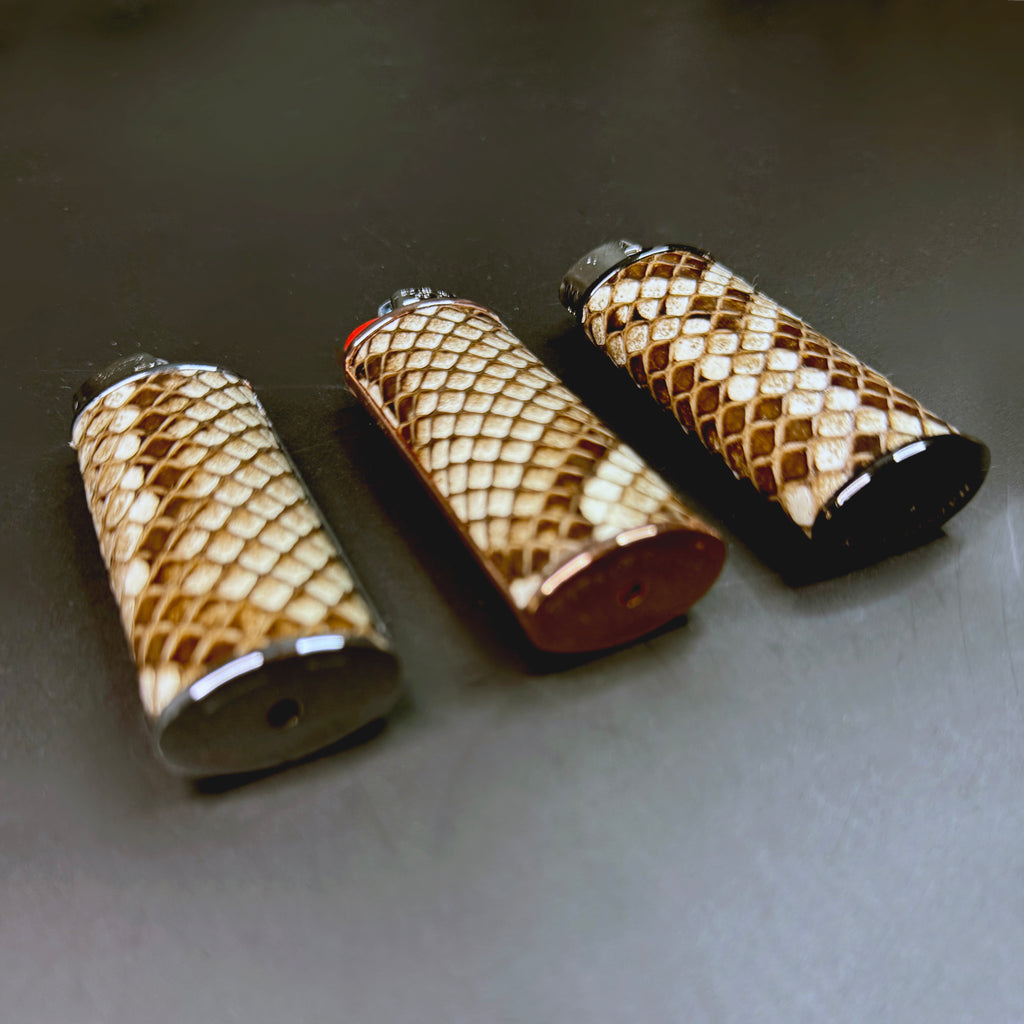 Angled close-up of three Burmese natural python lighter cases lying flat, highlighting the rich texture, color variations, and metallic bases that reflect light against a dark surface.