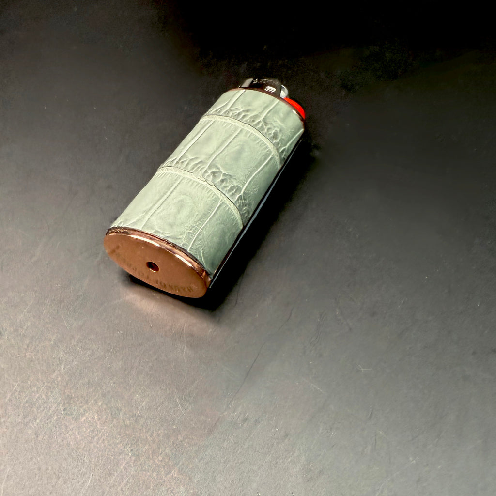 Angled view of the grey crocodile leather lighter lying on a dark surface, highlighting the texture of the leather and the reflective rose gold base.