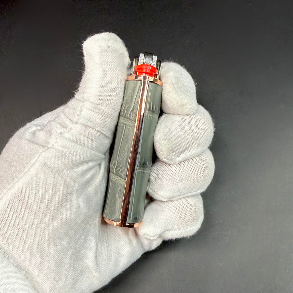 Close-up of a luxury lighter wrapped in genuine grey crocodile leather with a rose gold base, held in a gloved hand against a dark background.