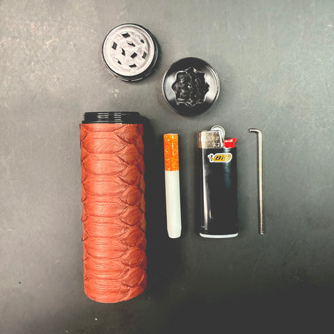 Disassembled view of all dugout components laid out on a dark surface — matte Syrah python case, grinder top and bottom, one-hitter, lighter, and metal poker tool.