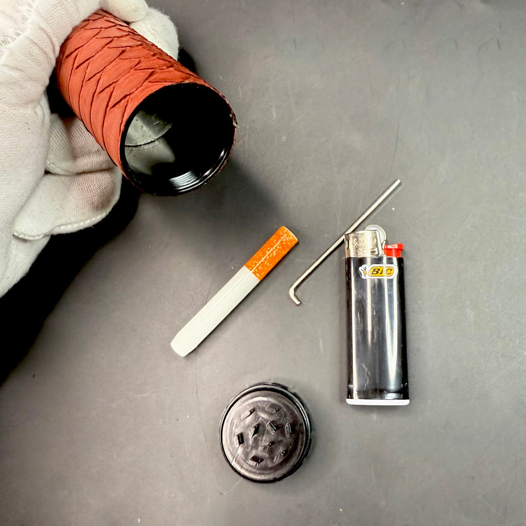 Close-up of a hand holding the dugout with the lid removed, displaying the interior cavity and disassembled components — one-hitter, lighter, poker tool, and grinder top.