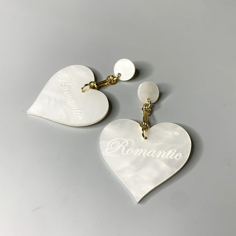 Oversized white acrylic heart earrings with "Romantic" engraving and gold chain detailing on black ear display
