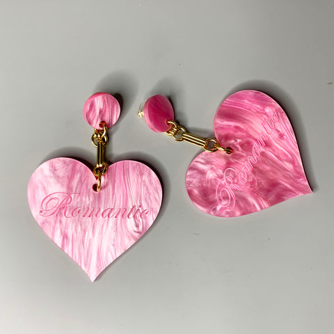 Oversized pink acrylic heart earrings with "Romantic" engraving and gold chain detailing on black ear display